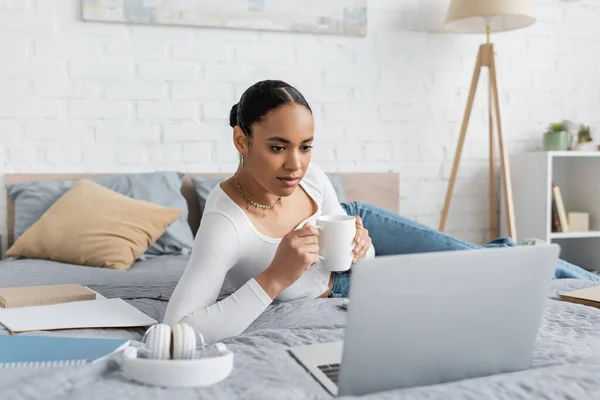 Junge afrikanisch-amerikanische Studentin hält Tasse Kaffee in der Hand und schaut auf Laptop im modernen Schlafzimmer — Stockfoto
