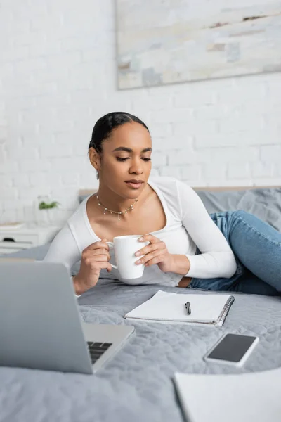 Young african american student holding cup of coffee and looking at notebook near laptop in modern bedroom — Stock Photo