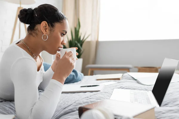Estudante americano africano bonita segurando xícara de café e olhando para laptop com tela em branco — Fotografia de Stock