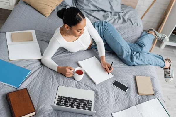 Draufsicht auf afrikanisch-amerikanische Studentin, die eine Tasse Tee hält und im Schlafzimmer auf Notizbuch schreibt — Stockfoto