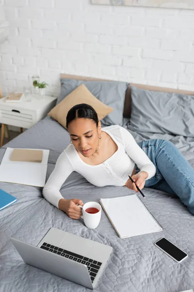 African american student holding cup of tea near laptop and notebook on bed at home — Stock Photo