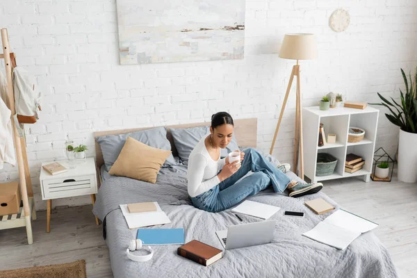 Étudiant afro-américain tenant une tasse de thé près des gadgets et des livres sur le lit à la maison — Photo de stock