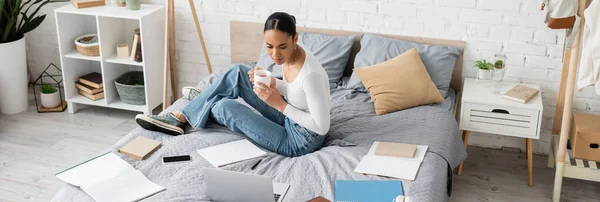 African american student holding tea near devices and notebooks on bed in bedroom, banner — Stock Photo