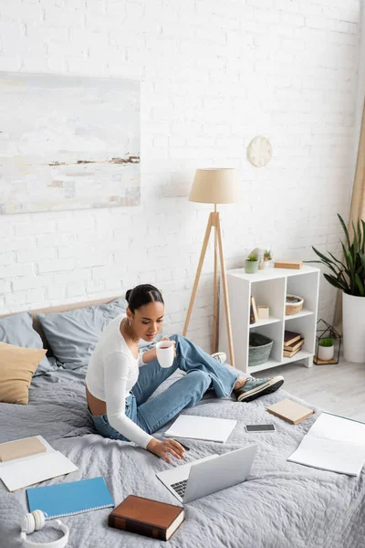 African american student holding tea and using laptop near notebooks and headphones on bed — Stock Photo