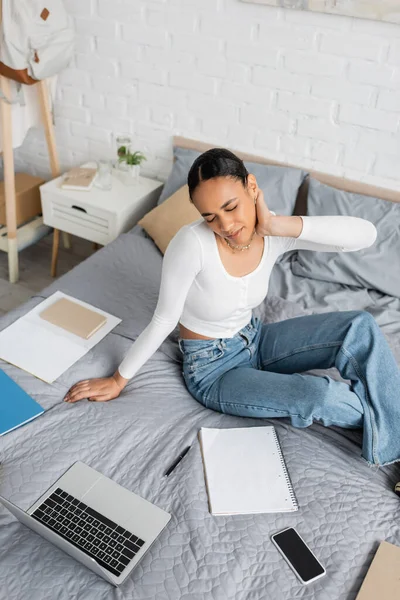 Уставший афроамериканец сидит рядом с ноутбуком и блокнотами на кровати — стоковое фото