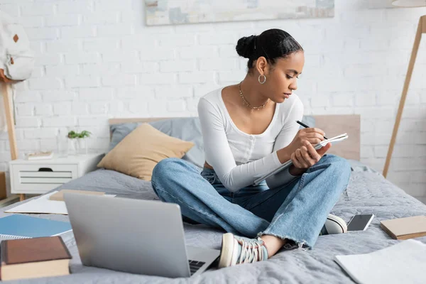 African american student writing on notebook near devices on bed at home — Stock Photo