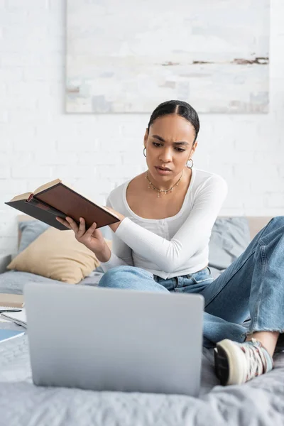 Konzentrierte afrikanisch-amerikanische Studentin hält Buch in der Hand und schaut zu Hause auf Laptop im Bett — Stockfoto