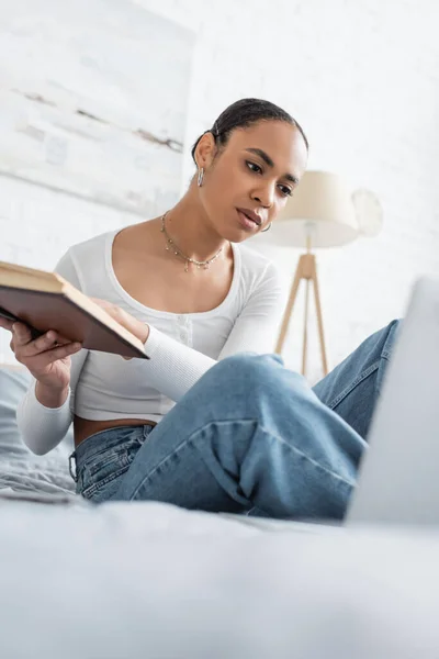 Африканський студент - американець тримає книжку біля розмитого ноутбука на ліжку вдома. — стокове фото