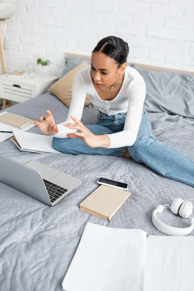 Estudante afro-americano conversando durante webinar no laptop perto de livros na cama em casa — Fotografia de Stock