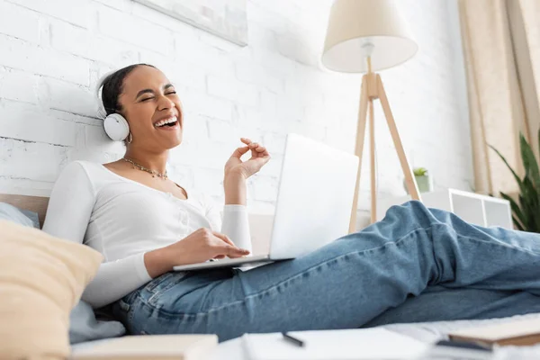 Cheerful african american student in headphones using laptop on bed at home — Stock Photo