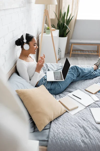 Side view of smiling african american student in headphones having video call on laptop on bed — Stock Photo