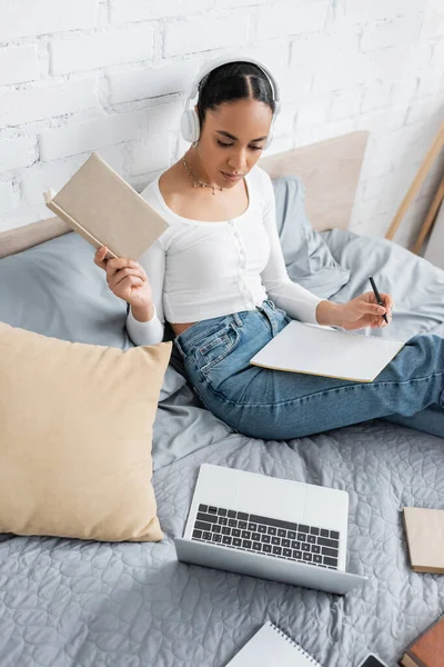African american student in headphones holding book near laptop and notebook on bed — Stock Photo
