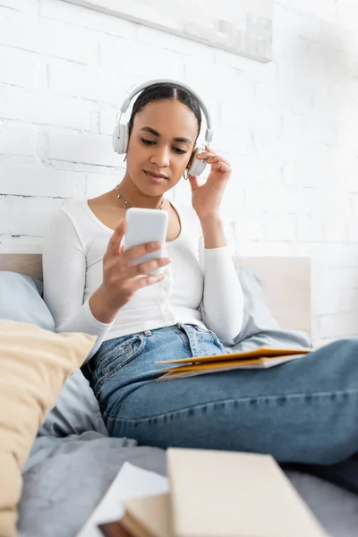 African american student in headphones using smartphone near books on bed — Stock Photo