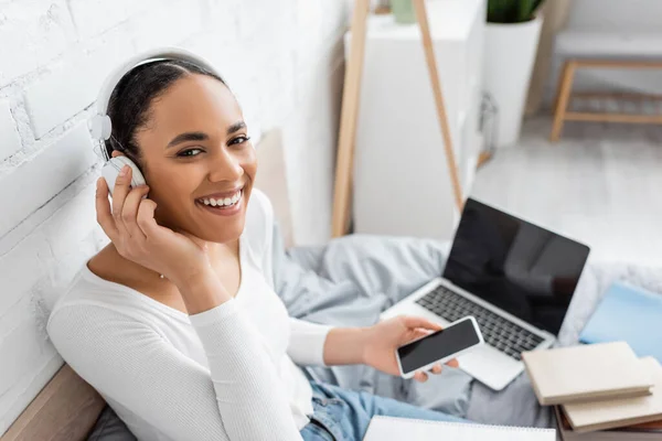 Cheerful african american student in headphones looking at camera near devices and book on bed — Stock Photo