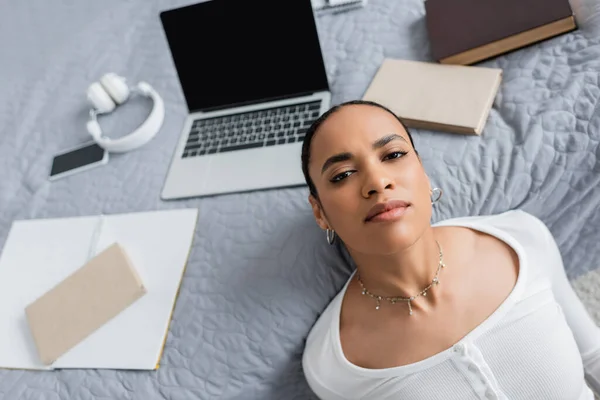Top view of african american student looking at camera near devices and books on bed — Stock Photo