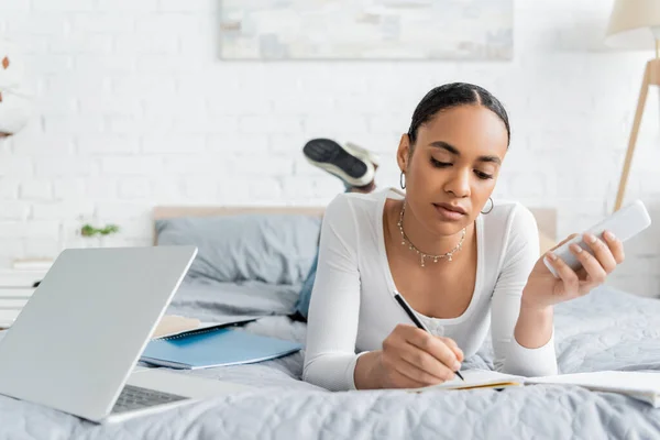 African american student holding smartphone and writing on notebook near laptop on bed — Stock Photo