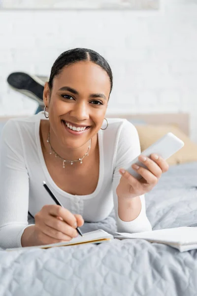 Positive african american student holding cellphone and writing on notebook on bed — Stock Photo