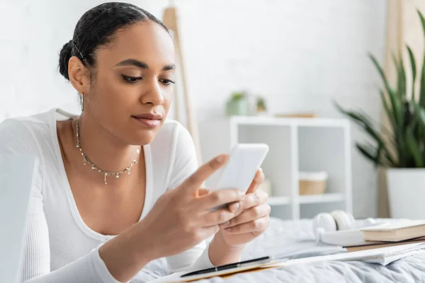 Smiling african american student using smartphone near blurred books on bed — Stock Photo