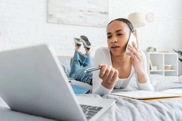 African american student talking on smartphone and looking at blurred laptop while lying on bed — Stock Photo