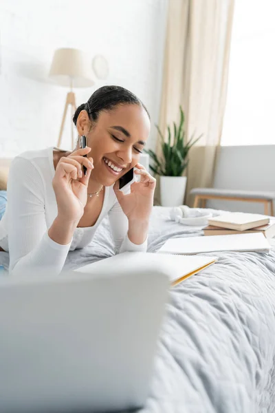 Cheerful african american student talking on cellphone near notebook and laptop on bed — Stock Photo