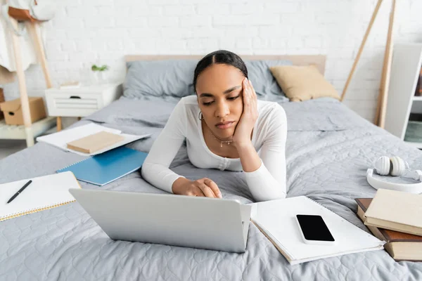 Étudiant afro-américain fatigué utilisant un ordinateur portable près des ordinateurs portables et smartphone sur le lit — Photo de stock