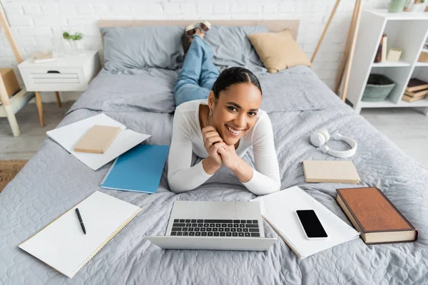 Glückliche afrikanisch-amerikanische Studentin liegt neben Geräten und Büchern auf dem Bett — Stockfoto
