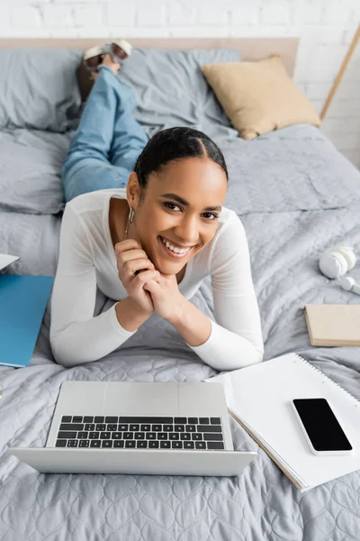 Cheerful african american student lying near gadgets and notebooks on bed — Stock Photo