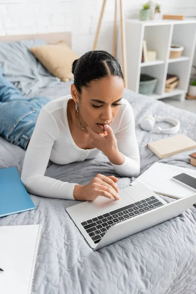 Étudiant afro-américain concentré utilisant un ordinateur portable près des ordinateurs portables sur le lit — Photo de stock