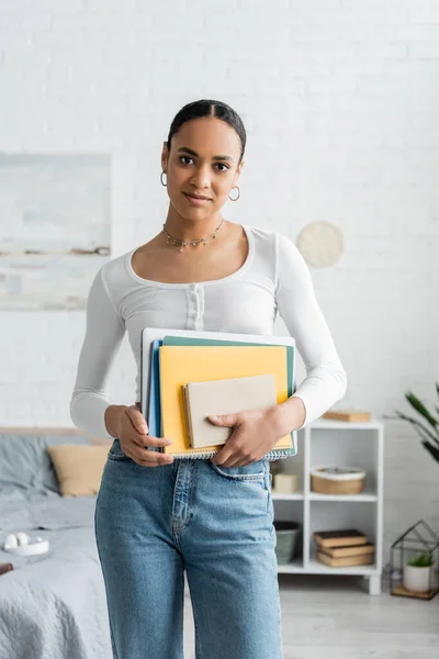 African american student holding notebooks and looking at camera in bedroom — Stock Photo