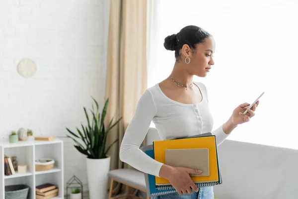 Side view of african american student using smartphone and holding notebooks at home — Stock Photo