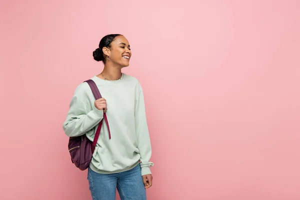 Alegre estudiante afroamericano con mochila mirando hacia otro lado aislado en rosa - foto de stock