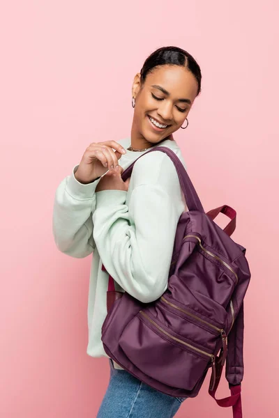 Молодой афроамериканец в толстовке смотрит на рюкзак, изолированный на розовом — стоковое фото