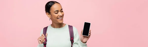 Усміхнений афроамериканський студент з рюкзаком тримає смартфон з порожнім екраном ізольовано на рожевому, банер — стокове фото