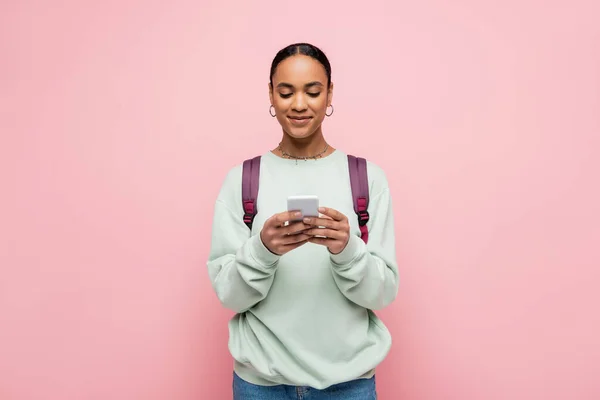 Smiling african american student with backpack typing message on smartphone isolated on pink — Stock Photo