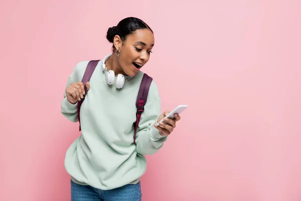 Emocionado estudiante afroamericano con auriculares usando teléfono inteligente aislado en rosa - foto de stock