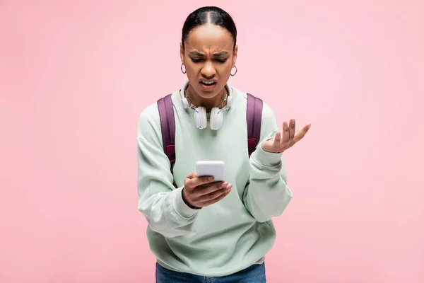 Angry african american student with headphones using mobile phone isolated on pink — Stock Photo