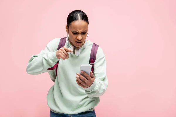 Angry african american student with backpack and headphones pointing at smartphone isolated on pink — Stock Photo