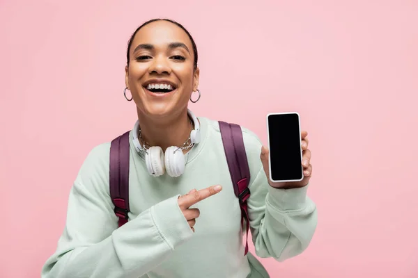 Estudiante afroamericano positivo con auriculares apuntando al teléfono celular con pantalla en blanco aislada en rosa - foto de stock