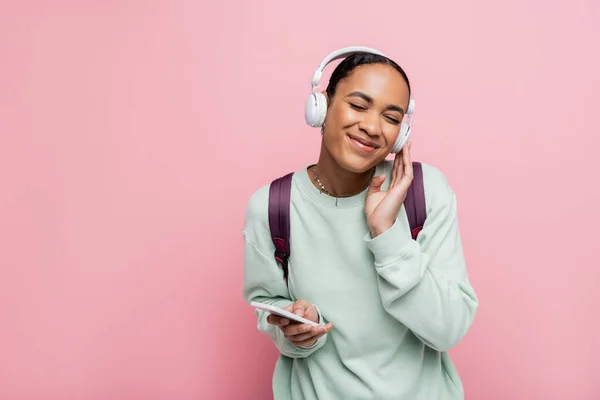Весела афроамериканка в бездротових навушниках тримає смартфон, насолоджуючись музикою ізольовано на рожевому — стокове фото