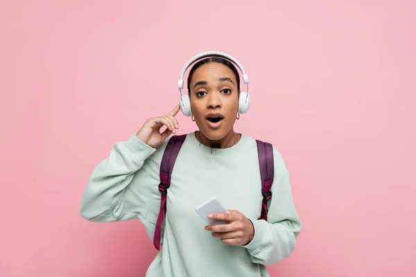 Asombrada mujer afroamericana en auriculares inalámbricos sosteniendo teléfono inteligente mientras escucha música aislada en rosa - foto de stock