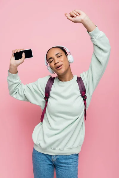 Joven estudiante afroamericano en auriculares inalámbricos sosteniendo el teléfono inteligente mientras escucha música y canta en rosa - foto de stock