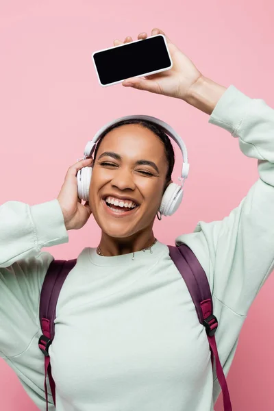 Felice donna afro-americana in cuffie wireless con smartphone mentre ascoltava musica isolata su rosa — Foto stock