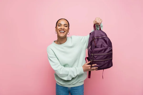 Happy african american student in sweatshirt holding purple backpack isolated on pink — Stock Photo