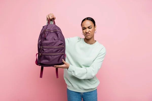 Displeased african american student in sweatshirt holding purple backpack isolated on pink — Stock Photo