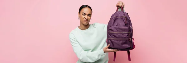 Étudiant afro-américain mécontent en sweat-shirt tenant sac à dos violet isolé sur rose, bannière — Photo de stock