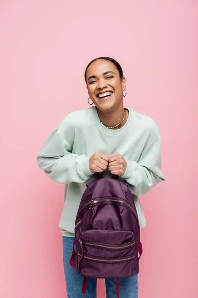 Joyful african american student in sweatshirt holding purple backpack isolated on pink — Stock Photo