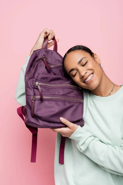 Задоволений афроамериканець з закритими очима, що тримає фіолетовий рюкзак ізольований на рожевому — стокове фото