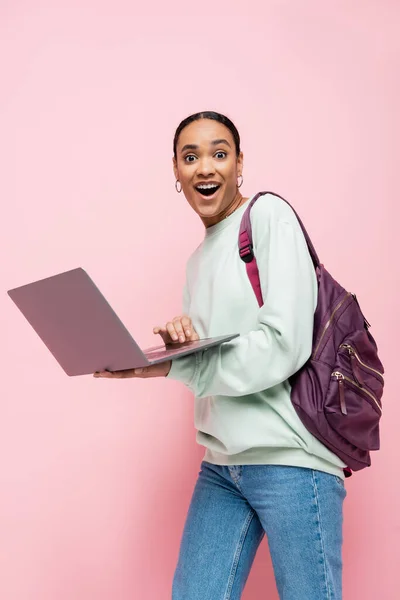 Erstaunt afrikanisch-amerikanischer Student mit Rucksack, Laptop in der Hand und Blick auf Kamera isoliert auf rosa — Stockfoto