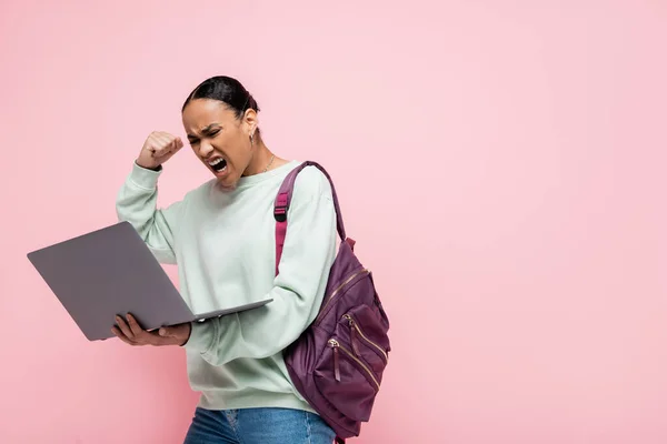 Раздраженный африканский американский студент с рюкзаком глядя на ноутбук изолирован на розовый — стоковое фото