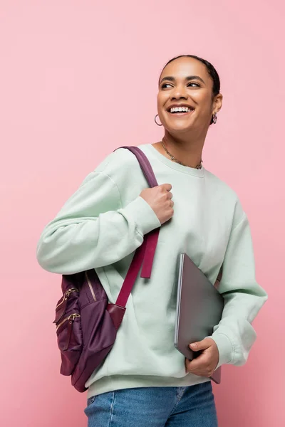 Joyful african american student with backpack holding laptop isolated on pink — Stock Photo